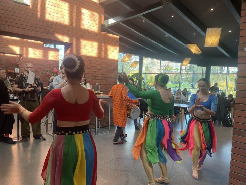 Trzy tancerki w kolorowych strojach tańczą, na dalszym planie tancerz oraz publiczność