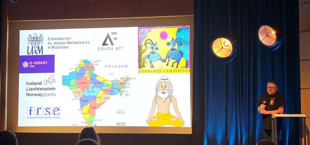 Profesor Krzysztof Stroński opowiada o platformie, wyświetla się slajd z logo UAM, GrupyAF, Funduszy Norweskich i Fundacji Rozwoju systemu Edukacji, ehindi. i Indologii UAM. Na slajdzie też mapa Indii w podziale administracyjnym i siedzący jogin.